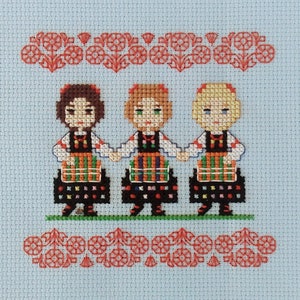Könnte beinhalten: Kreuzstich-Stickerei mit drei Mädchen in traditionellen Trachten, die sich an den Händen halten und lächeln. Die Mädchen tragen bunte Kleider und Schürzen, mit schwarzen Haaren und roten Akzenten. Der Hintergrund ist hellblau mit einem roten floralen Rand.