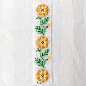 May include: A white cross-stitch bookmark featuring a floral design. The bookmark has three yellow and orange flowers with green leaves and stems. The background is a white fabric with vertical stripes.