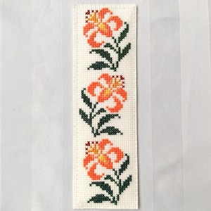Könnte beinhalten: Handgefertigter Lesezeichen mit einem Kreuzstich-Blumenmuster. Der cremefarbene Hintergrund zeigt drei orangefarbene und gelbe Lilien, jede mit grünen Blättern und Stielen. Die Stickerei ist detailliert und präzise.