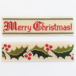 May include: Two white canvas panels with cross-stitch embroidery. The top panel has the words "Merry Christmas!" in red thread. The bottom panel has a green holly leaf design with red berries.