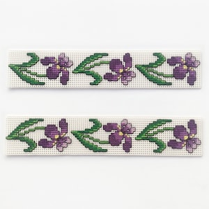 May include: Two white fabric strips with a purple and green floral embroidery design. The design features a repeating pattern of purple flowers with yellow centers and green leaves.
