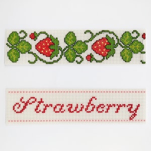 May include: Two white cross-stitch pieces with red embroidery. The top piece features a green vine with red strawberries. The bottom piece says "Strawberry" in red.