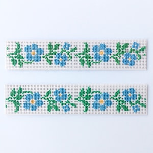 May include: Two white canvas strips with blue and green embroidered flowers. The flowers are a simple design with five petals and a yellow center.