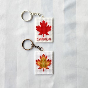 May include: Two rectangular keychains featuring cross-stitch designs on white fabric. The top keychain displays a red maple leaf with the word CANADA stitched in red. The bottom keychain features a multi-coloured autumn maple leaf in red, orange, and yellow.