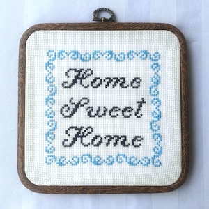 May include: A square cross-stitch embroidery with the words "Home Sweet Home" in black thread. The text is centered on a white background, framed by a blue border and a wooden frame with a metal loop for hanging.