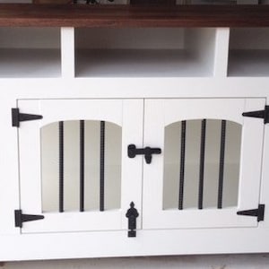 May include: White wooden dog crate with two doors and a top shelf. The crate has black metal bars and black metal hinges. The doors have a black metal latch.