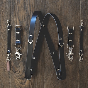 May include: Black leather camera straps with silver hardware and metal clips. The straps are crossed and laid out on a wooden surface.