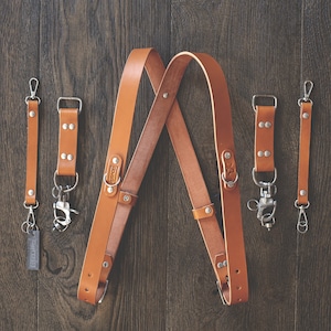 May include: A set of brown leather camera straps with silver hardware. The straps are designed to be worn across the body and have adjustable lengths. The straps are made from high-quality leather and are durable and comfortable to wear.