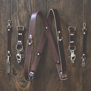 May include: A set of four brown leather camera straps with silver hardware. The straps are arranged in a criss-cross pattern on a wooden surface.