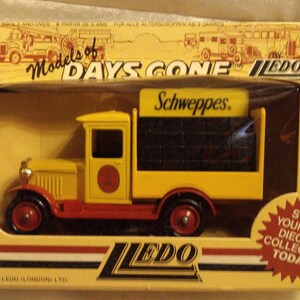 May include: A yellow and red diecast model truck with the Schweppes logo on the side. The truck is in its original box, which features the "Models of Days Gone" logo and the Lledo brand.