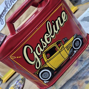 Könnte beinhalten: Ein roter Metall-Benzinkanister mit einer gelben Hot-Rod-Illustration und dem Wort "Gasoline" in einer stilisierten Schriftart. Der Kanister hat einen Griff und einen Ausguss, zum Aufbewahren von Kraftstoff.