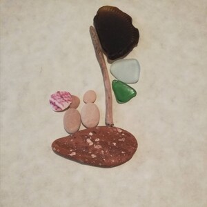 Könnte beinhalten: Ein Steinbild mit braunem, grünem und weißem Meerglas, einer rosa-weißen Muschel und braunem Treibholz auf einem braunen Stein.