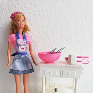 May include: A Barbie doll wearing a pink shirt, blue and white striped apron, and a red and white bandana. She is standing in front of a white play kitchen with a pink mixing bowl, two silver utensils, and two clear measuring cups.