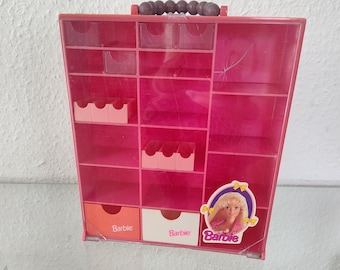 Puede incluir: Caja de almacenamiento de plástico rosa con múltiples compartimentos y cajones. La caja tiene un asa y un logotipo de Barbie con una imagen de Barbie en la parte delantera. Los cajones están etiquetados con "Barbie".