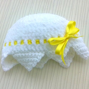 May include: White crocheted baby hat with a yellow satin bow. The hat has a scalloped edge and a row of yellow squares around the brim.