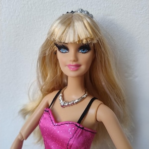 May include: A doll with blonde hair, blue eyes, and a pink dress with black accents. The doll is wearing a silver necklace and a tiara. The dress has a sparkly texture.