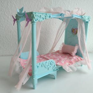 Może przedstawiać: Miniaturowe łóżko dla lalek w jasnoniebieskim kolorze z baldachimem i różowymi akcentami. Łóżko ma różowo-biały koc w kształcie serca, różową poduszkę i przezroczyste różowe zasłony. Rama łóżka ma dekoracyjne detale kwiatowe.