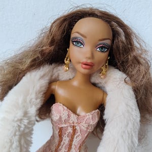 Könnte beinhalten: Modepuppe mit braunen Haaren, blauen Augen und goldenen Ohrringen. Die Puppe trägt ein rosa Spitzenkorsett und eine weiße Kunstfellstola. Das Make-up der Puppe umfasst rosa Lippenstift und Lidschatten. Sammlerstück.