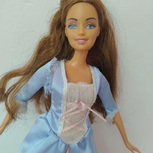 以下が含まれることがあります： 長い茶色の髪と青い目をした人形が、水色のサテンのドレスを着ています。ドレスには、ピンクのリボンの編み上げディテールが施された白いボディスパネル、ネックラインに沿ったシルバーのトリム、そして端がほつれた水色の袖が付いています。