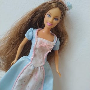 以下が含まれることがあります： 長い茶色の髪、青い目、ピンクの口紅をした人形。 人形は、ピンクと白のボディスと、青とピンクのアクセントが付いたお揃いのヘッドバンドが付いた、水色のドレスを着ています。 ドレスはキラキラしたデザインです。