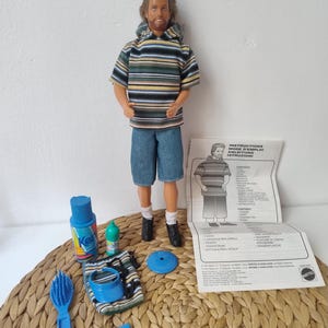 Könnte beinhalten: Eine männliche Puppe mit braunem Haar und Bart, bekleidet mit einem gestreiften Kapuzenpullover und blauen Jeansshorts. Das Zubehör umfasst eine blaue Haarbürste, einen blauen Rasierer, ein blaues Visier, eine blaue Sprühflasche, eine grüne Flasche und eine Anleitung.
