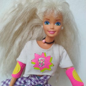May include: A vintage doll with long, white, fluffy hair. She wears a white crop top with a pink and green star design, blue jeans, and pink armbands with yellow accents. A purple and white checkered shirt is tied around her waist.