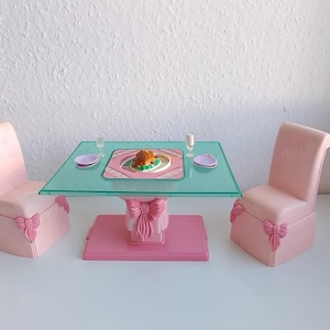 May include: A miniature pink dining set with a glass tabletop, two chairs, and a table setting. The chairs have bow details. The table is set with a plate of food, two glasses, and two small plates. The set is made of plastic.