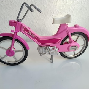 Könnte beinhalten: Ein rosa Spielzeug-Moped mit weißem Sitz, grauer Lenkstange und schwarzen Reifen. Der Rahmen ist mit einer rosa-weißen Grafik und dem Schriftzug Barbie versehen. Das Fahrzeug verfügt über ein silberfarbenes Motordetail und einen Ständer.