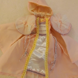 Könnte beinhalten: Ein rosa Puppenkleid mit einem weißen Satin-Mieder, gelbem Besatz und einem passenden rosa Kapuzenumhang mit Spitzenbesatz.