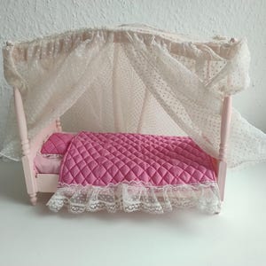 May include: A miniature pink canopy bed with a quilted pink blanket and pillow. The bed frame is light pink, and the canopy is made of white dotted fabric. The bed has white lace trim.