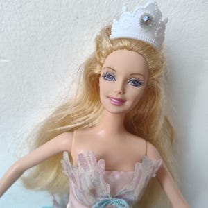 Puede incluir: Muñeca con pelo largo y rubio, que lleva una corona blanca con una gema. La muñeca viste un corpiño rosa con un lazo azul y una falda azul y blanca. Los brazos de la muñeca están extendidos.