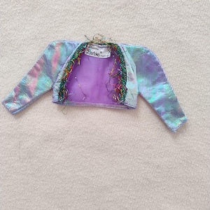 Könnte beinhalten: Eine lilafarbene, irisierende Barbie Fashion-Puppenjacke mit langen Ärmeln und einem glitzernden Besatz. Die Jacke hat eine holografische Oberfläche und ist aus einem durchsichtigen Stoff gefertigt.