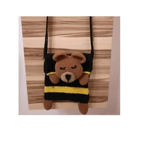 Könnte beinhalten: Eine schwarze und gelbe gestreifte gehäkelte Tasche mit einem braunen gehäkelten Bärenkopf auf der Vorderseite. Die Tasche hat einen langen Riemen zum Tragen über der Schulter.