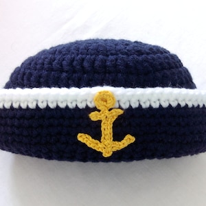 Può includere: Un cappello da marinaio in crochet blu con una fascia bianca e un emblema di ancora giallo.