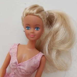 Puede incluir: Muñeca vintage con cabello rubio recogido en una cola de caballo. La muñeca tiene ojos azules y viste un vestido rosa con escote en V. El vestido tiene un aspecto texturizado y brillante. La muñeca está sobre un fondo blanco liso.