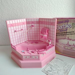 Könnte beinhalten: Ein rosa Barbie-Badezimmer-Set mit einer sechseckigen Badewanne, Dusche und Zubehör. Die Wände sind mit rosa Quadraten gefliest. Eine Schachtel mit dem Barbie-Logo ist sichtbar, zusammen mit einem Anleitungsblatt.
