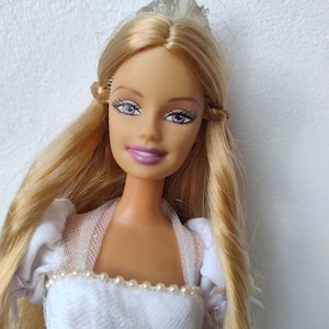 Pode incluir: Uma boneca loira com tiara e vestido branco. A boneca tem cabelo longo e esvoaçante e usa um vestido branco com detalhes de pérolas. O rosto da boneca tem maquiagem, incluindo sombra roxa e batom rosa.