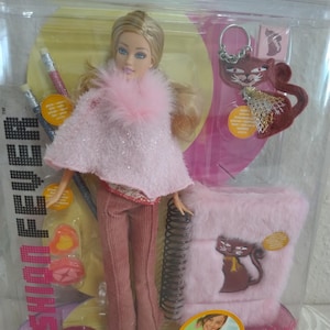 Peut inclure: Poupée mannequin dans un étui en plastique transparent. La poupée a les cheveux blonds et porte une tenue rose avec un col en fausse fourrure. Les accessoires comprennent un carnet rose, des crayons et un porte-clés en forme de chat. L'emballage porte les mots "Fashion Fever".