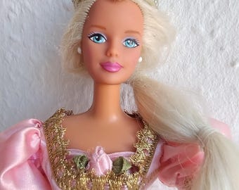 May include: A vintage doll with blonde hair, blue eyes, and a pink dress. The doll wears a silver tiara and has a long ponytail. The dress features gold trim and a corset-style bodice, with a pink and gold colour scheme.