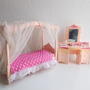 May include: A miniature dollhouse bedroom set with a pink canopy bed, a quilted pink comforter, and a matching vanity with a mirror. The bed has a white lace trim and sheer white fabric. The vanity has drawers and a mirror.
