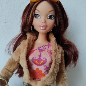 Könnte beinhalten: Eine Modepuppe mit rotbraunem Haar, die eine hellbraune Kunstfelljacke, ein pink-orange gemustertes Oberteil und eine blaue Jeans trägt. Die Puppe hat eine Sonnenbrille auf dem Kopf und silberne Creolen. Eine goldfarbene Handtasche ist sichtbar.
