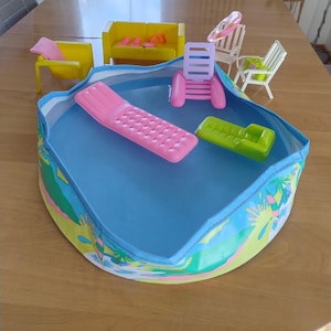 以下が含まれることがあります： 青い内装とカラフルな熱帯の縁取りが施された、ミニチュアのドールハウスのプール。 セットには、ピンクのプールフロート、緑のラウンジチェア、小さな黄色と白の家具が含まれています。
