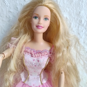 以下が含まれることがあります： 緑色の目とピンクのドレスを着たブロンドの人形。人形のドレスは、ピンクのアクセントと金色のトリムが付いた白いレースのボディスが特徴です。スカートは薄いピンクのチュールです。人形は長く、流れるようなブロンドの髪をしています。