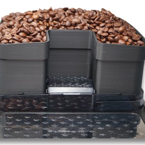 Puede incluir: Un tolva de granos de café negro con dos compartimentos separados para diferentes tipos de granos. La tolva está llena de granos de café.