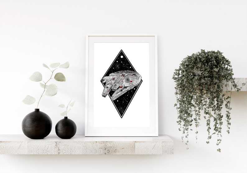 May include: A framed print featuring a detailed illustration of the Millennium Falcon starship. The artwork is set against a black diamond shape with a starry background. The print is displayed on a white shelf with decorative black vases and greenery.