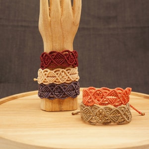 Könnte beinhalten: Vier Makramee-Armbänder in Rot-, Beige- und Lilatönen werden auf einer Holzhand präsentiert. Ein fünftes orangefarbenes und ein sechstes beigefarbenes Armband befinden sich auf einem Holztablett. Die Armbänder haben ein gewebtes Design.