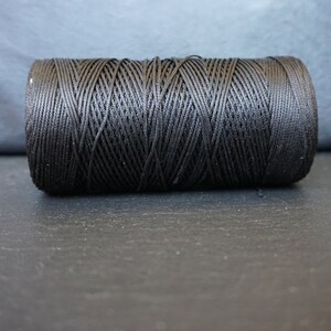 May include: A spool of black thread, tightly wound and cylindrical in shape. The thread is a deep, solid black, and the texture of the wound thread is clearly visible. The spool is resting on a dark surface.