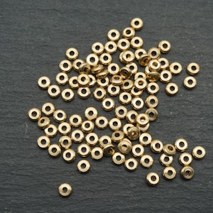 Könnte beinhalten: Eine Ansammlung kleiner, goldfarbener Metallperlen mit kreisförmiger Form und einem Loch in der Mitte. Die Perlen sind auf einer dunkelgrauen, strukturierten Oberfläche verstreut, was einen Kontrast zwischen den glänzenden Perlen und dem matten Hintergrund erzeugt.