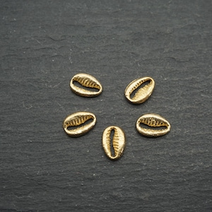 Könnte beinhalten: Fünf kleine, goldfarbene Kaurischalen-Anhänger auf einer dunklen, strukturierten Oberfläche angeordnet. Jede Muschel hat eine detaillierte, geriffelte Textur im Inneren. Die Muscheln sind gleichmäßig verteilt.