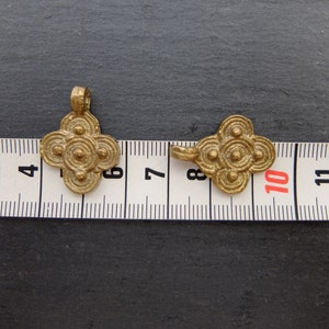 May include: Two antique-style, gold-colored pendants with a four-petal flower design. Each pendant features concentric circles and small spheres. The pendants are placed on a measuring tape, with one pendant measuring approximately 1.5 inches in length.